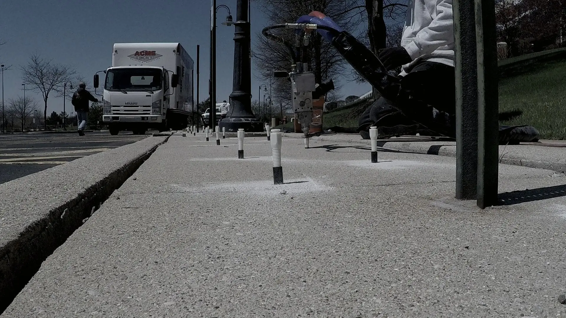 Low-angle view down a line of injection ports lifting a settled sidewalk, an Acme Concrete Raising truck parked alongside